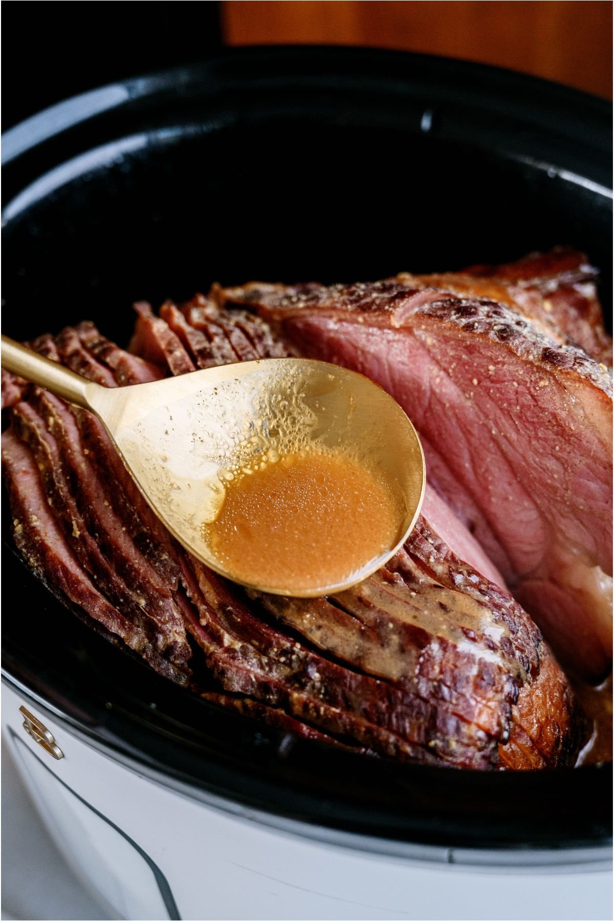 A ham in the crock pot with a serving spoon spooning maple glaze over ham.