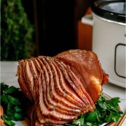 Crockpot Ham (With Maple Glaze) sliced on a serving plate with garnishes in front of a slow cooker.