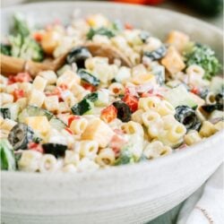 A white serving bowl full of creamy pasta salad. With bell peppers in the background.