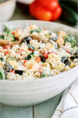 A white serving bowl full of creamy pasta salad. With bell peppers in the background.