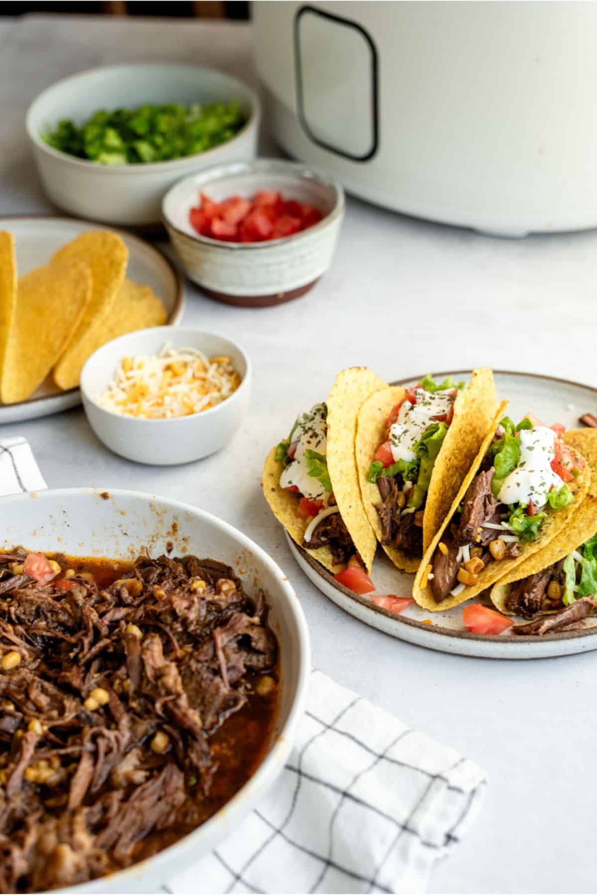 A plate of Slow Cooker Shredded Beef Tacos and ingredients needed to make Slow Cooker Shredded Beef Tacos
