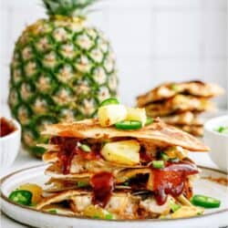 Stacked pineapple quesadilla topped with jalapeño slices on a plate; a whole pineapple in the background.