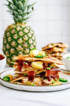 Stacked pineapple quesadilla topped with jalapeño slices on a plate; a whole pineapple in the background.