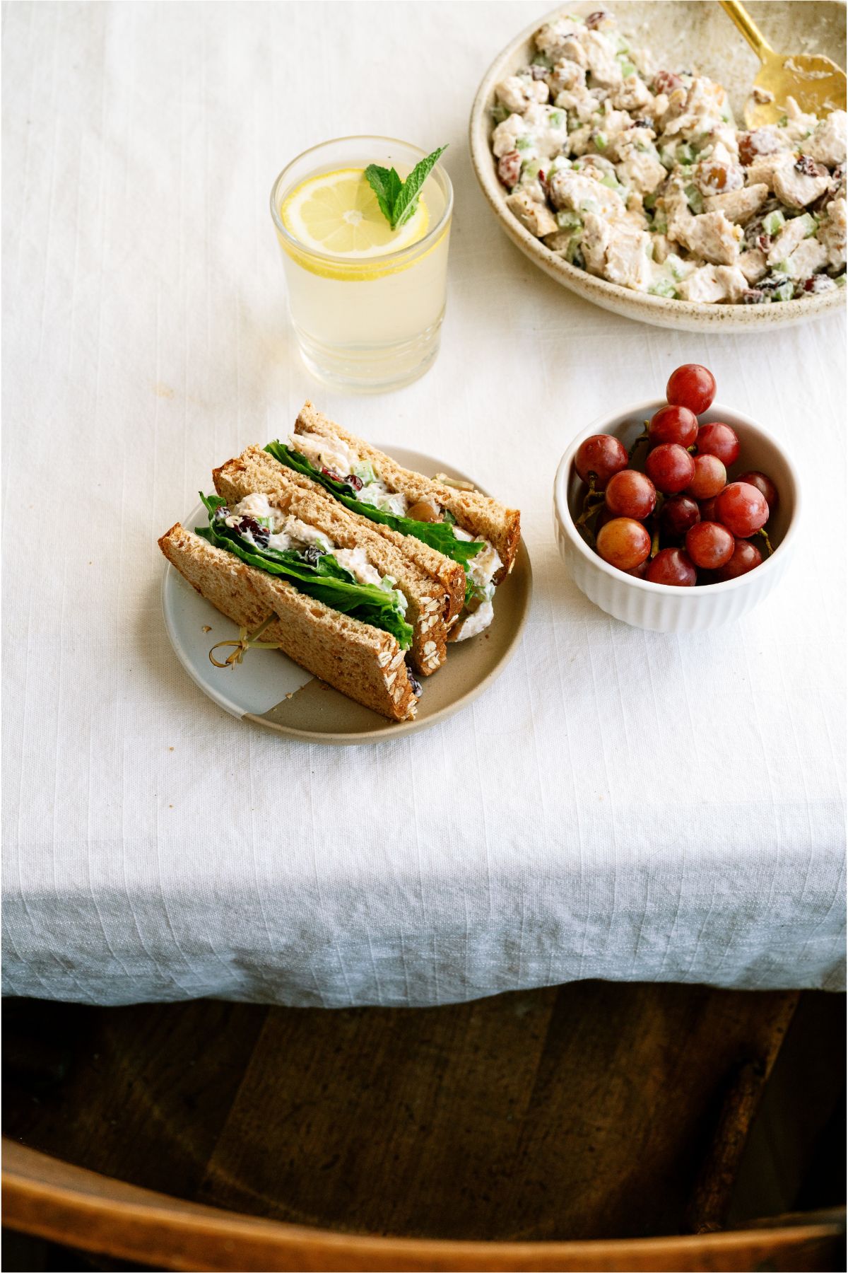 A bowl of Chicken Salad, a Chicken Salad Sandwiches cut in half on a plate with a small bowl of grapes and a glass of lemonade