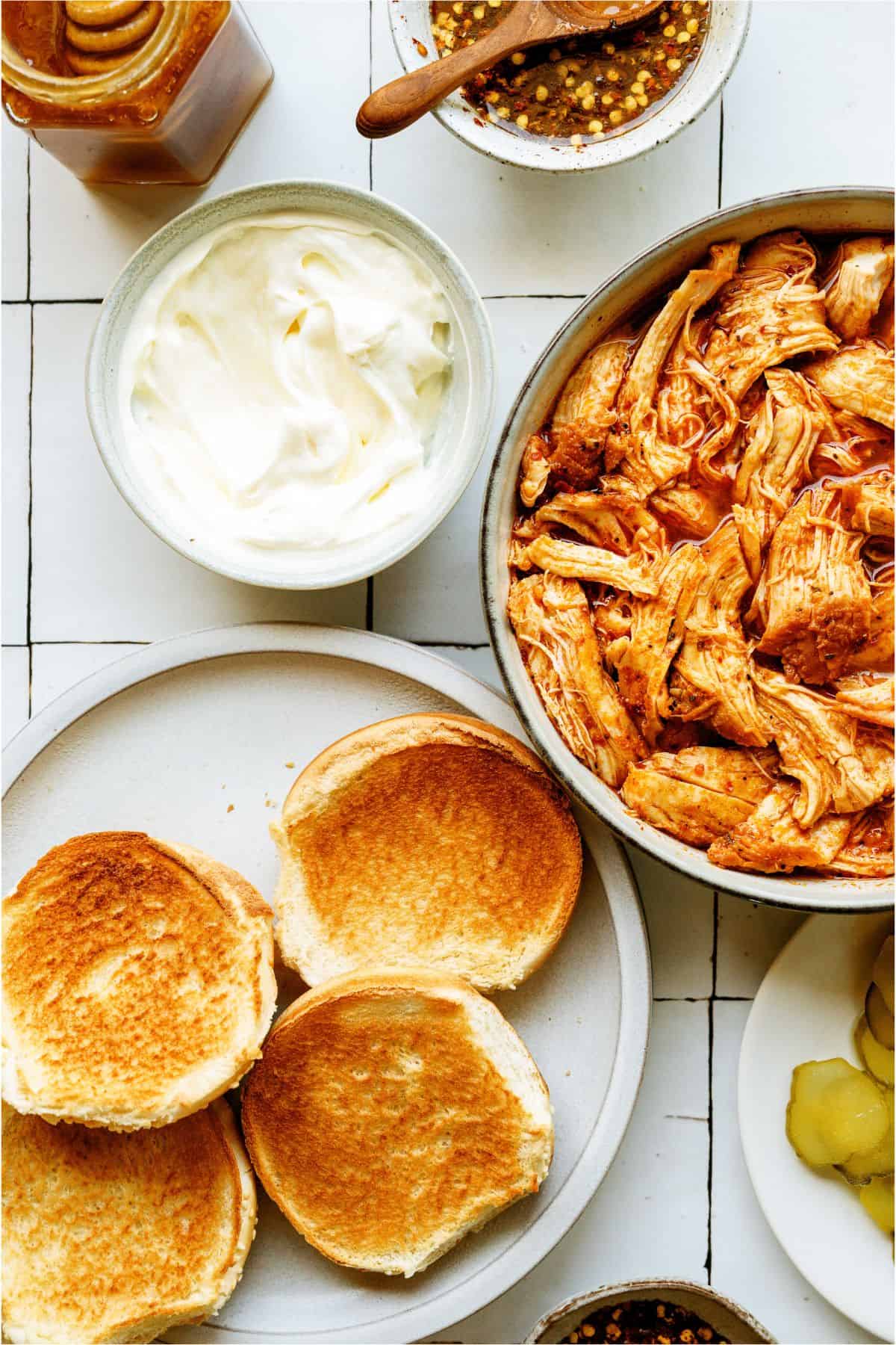 Instant Pot Hot Honey Chicken in a bowl, a small bowl of mayo, buns, a small bowl of hot honey