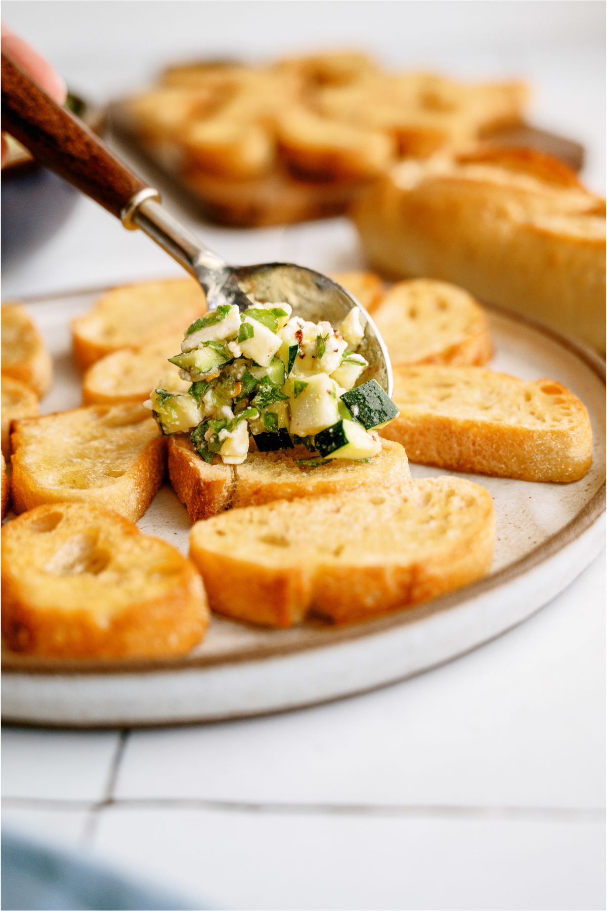 Spooning Zucchini Feta Bruschetta on top of sliced and toasted baguette bread 