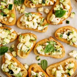 A plate of bruschetta topped with diced zucchini, crumbled feta cheese, red pepper flakes, and fresh basil leaves.