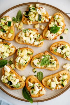 A plate of bruschetta topped with diced zucchini, crumbled feta cheese, red pepper flakes, and fresh basil leaves.