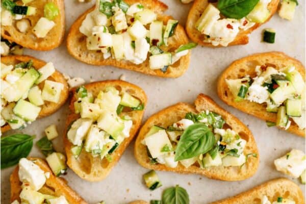 A plate of bruschetta topped with diced zucchini, crumbled feta cheese, red pepper flakes, and fresh basil leaves.