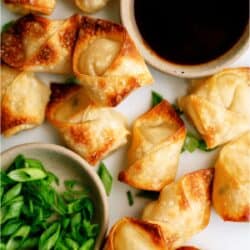 Fried wontons arranged next to bowls of dipping sauce and chopped green onions on a white surface.