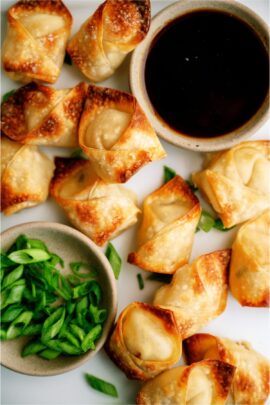 Fried wontons arranged next to bowls of dipping sauce and chopped green onions on a white surface.