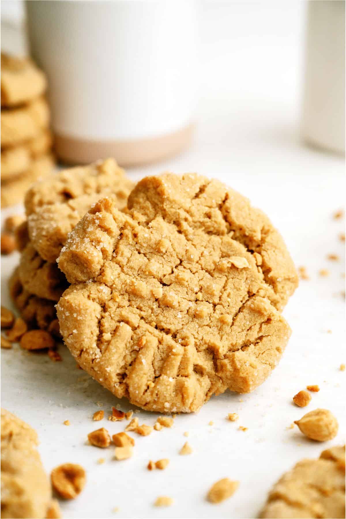 Peanut butter cookies stacked with a crumbly texture on a white surface, surrounded by small bits of peanuts.