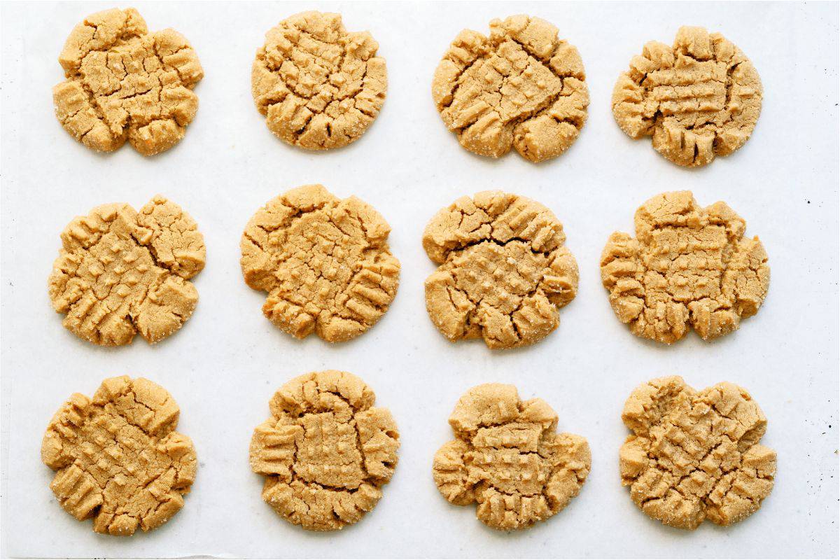 Twelve peanut butter cookies arranged in rows on a white surface, each with a crisscross pattern on top.