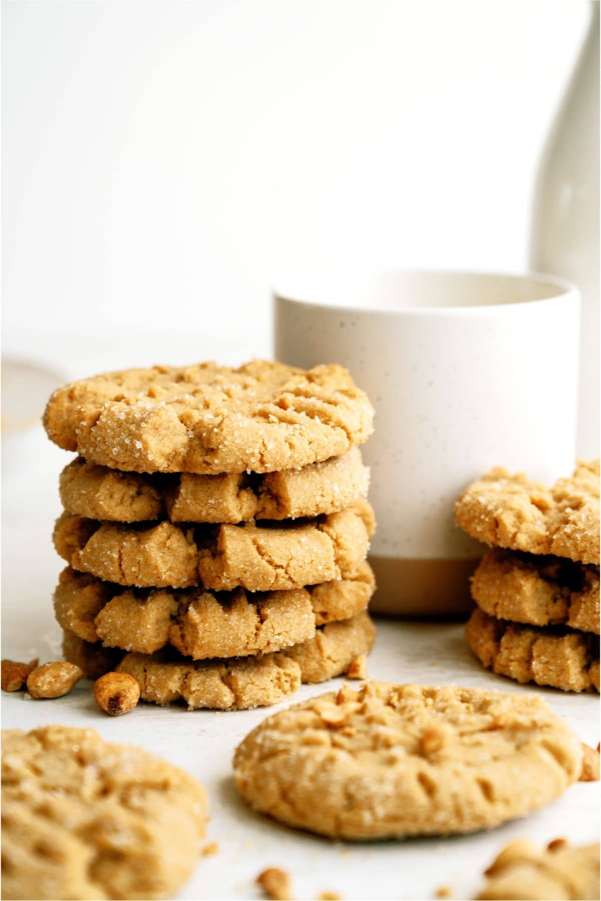 Stacked peanut butter cookies with sugar crystals sit next to a beige mug on a white surface.