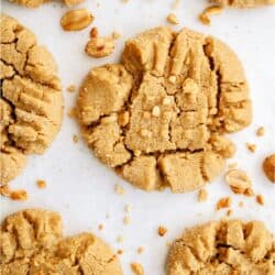 Close-up of six peanut butter cookies with a crisscross pattern on a white surface, surrounded by scattered peanuts.