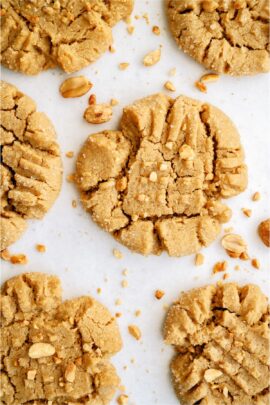 Close-up of six peanut butter cookies with a crisscross pattern on a white surface, surrounded by scattered peanuts.