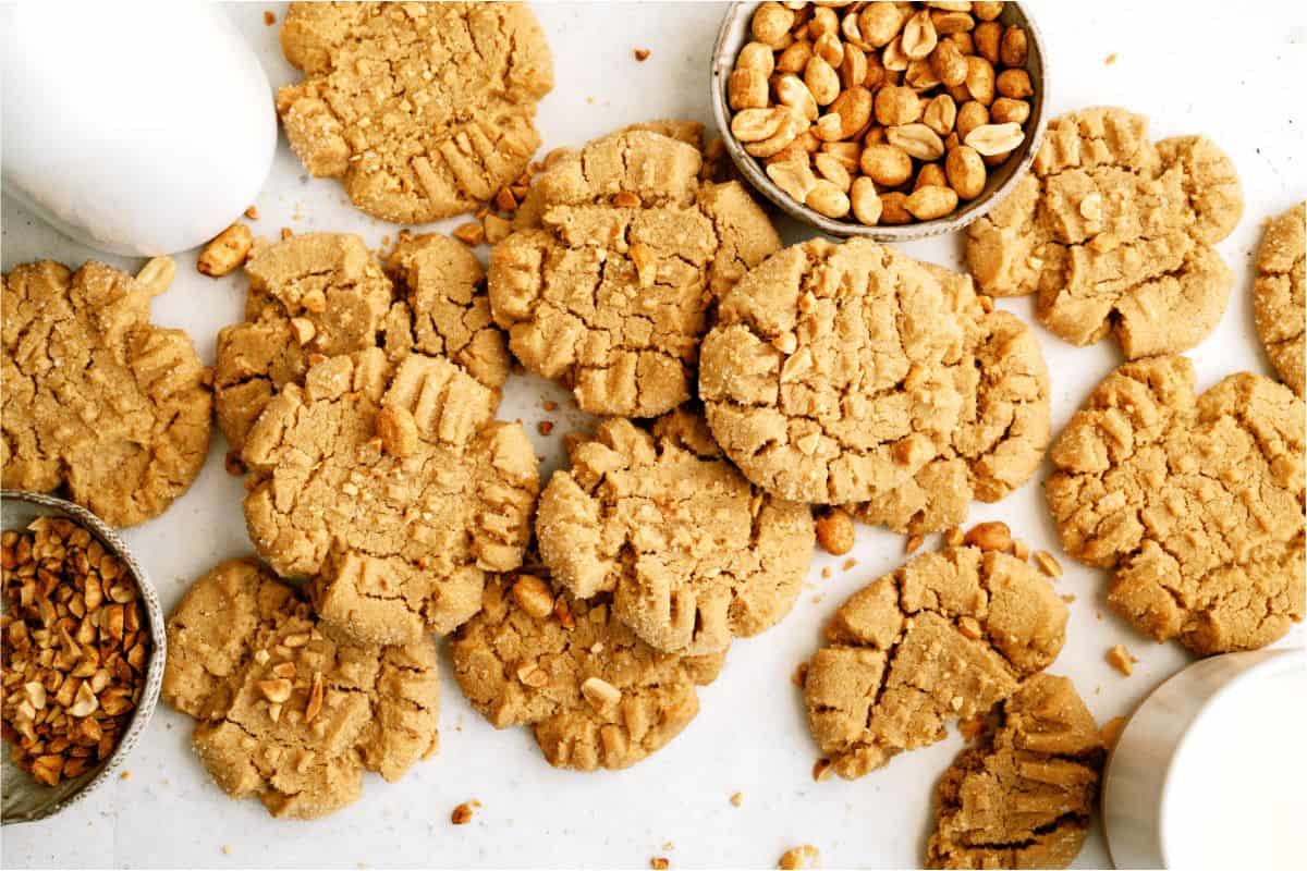 Several peanut butter cookies surrounded by bowls of peanuts and chopped nuts on a white surface.