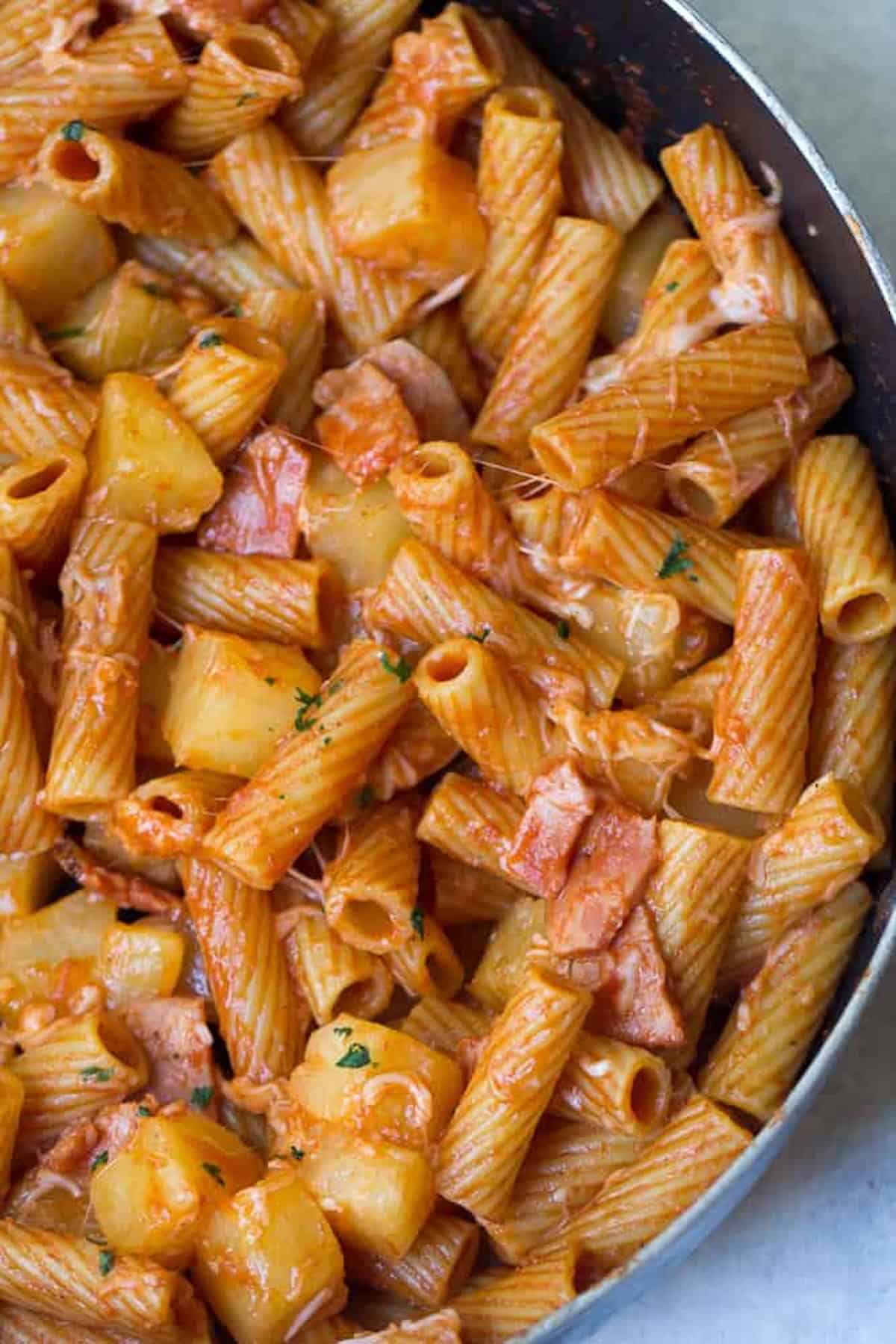 A skillet filled with rigatoni pasta in tomato sauce, mixed with chunks of potatoes and pieces of meat, garnished with bits of herbs.