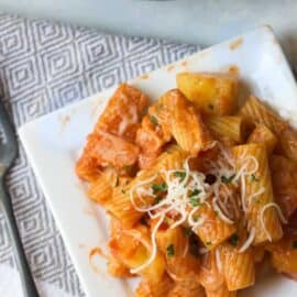 A square white plate with rigatoni pasta in tomato sauce, garnished with shredded cheese and herbs. A fork is placed nearby, and a pan with more pasta is partially visible in the background.