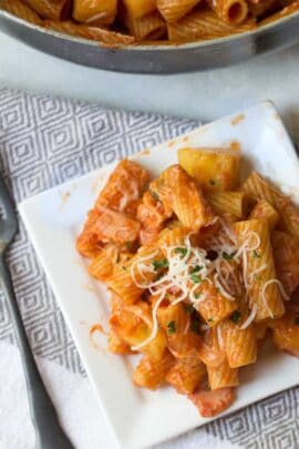 A square white plate with rigatoni pasta in tomato sauce, garnished with shredded cheese and herbs. A fork is placed nearby, and a pan with more pasta is partially visible in the background.