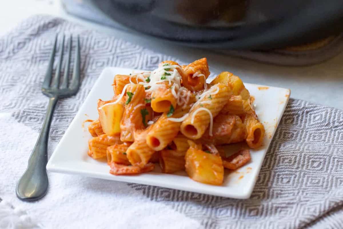A square white plate with a serving of rigatoni pasta topped with tomato sauce and grated cheese, placed on a patterned cloth napkin beside a fork.
