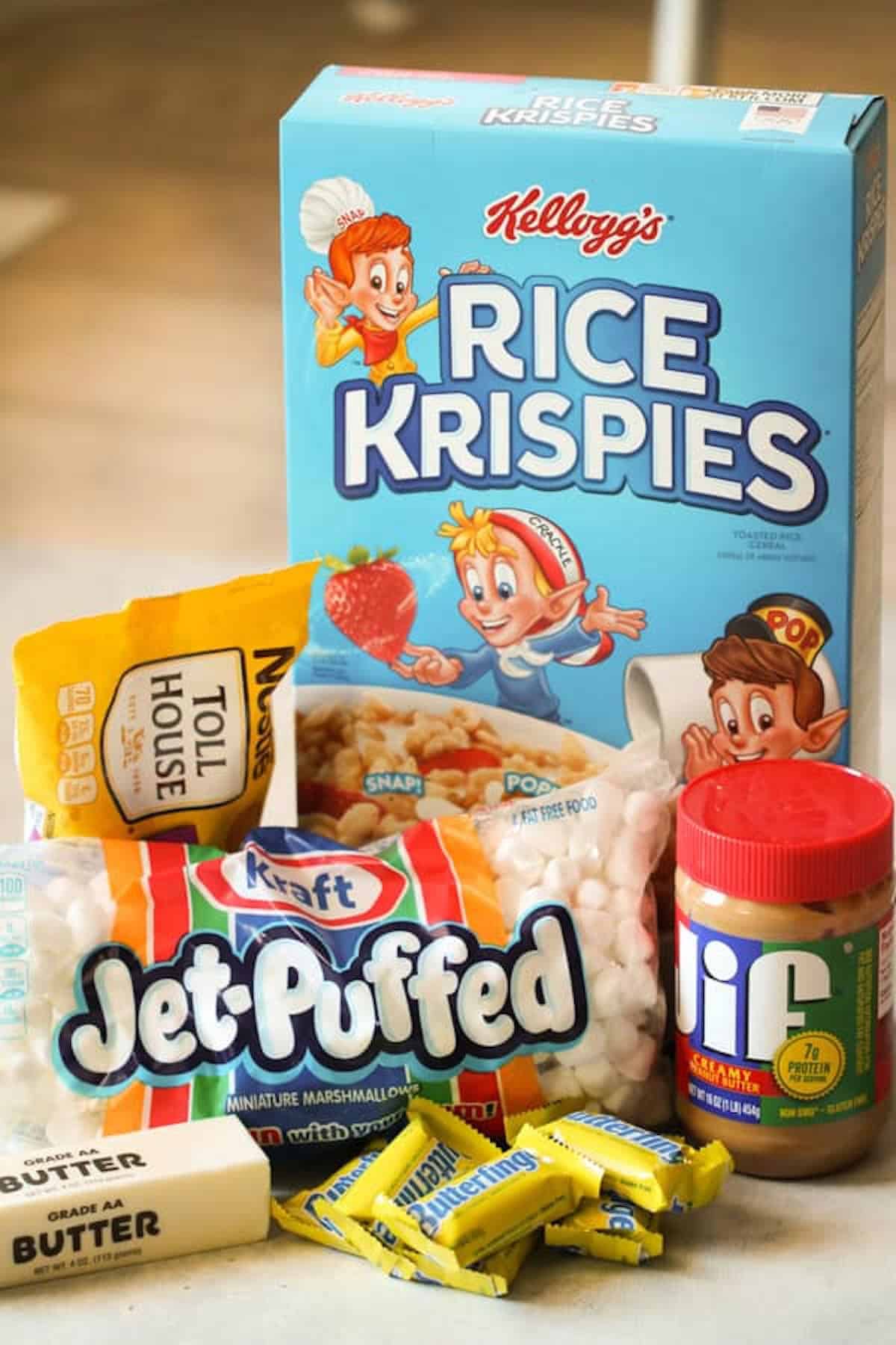 A box of Rice Krispies, a bag of Jet-Puffed marshmallows, a jar of Jif peanut butter, a pack of Toll House chocolate chips, a stick of butter, and several wrapped Butterfinger candies.