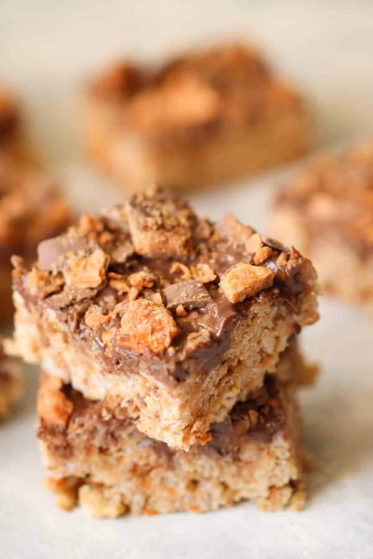 Two stacked, square-shaped rice crispy treats topped with crumbled toffee bits, with additional treats blurred in the background.