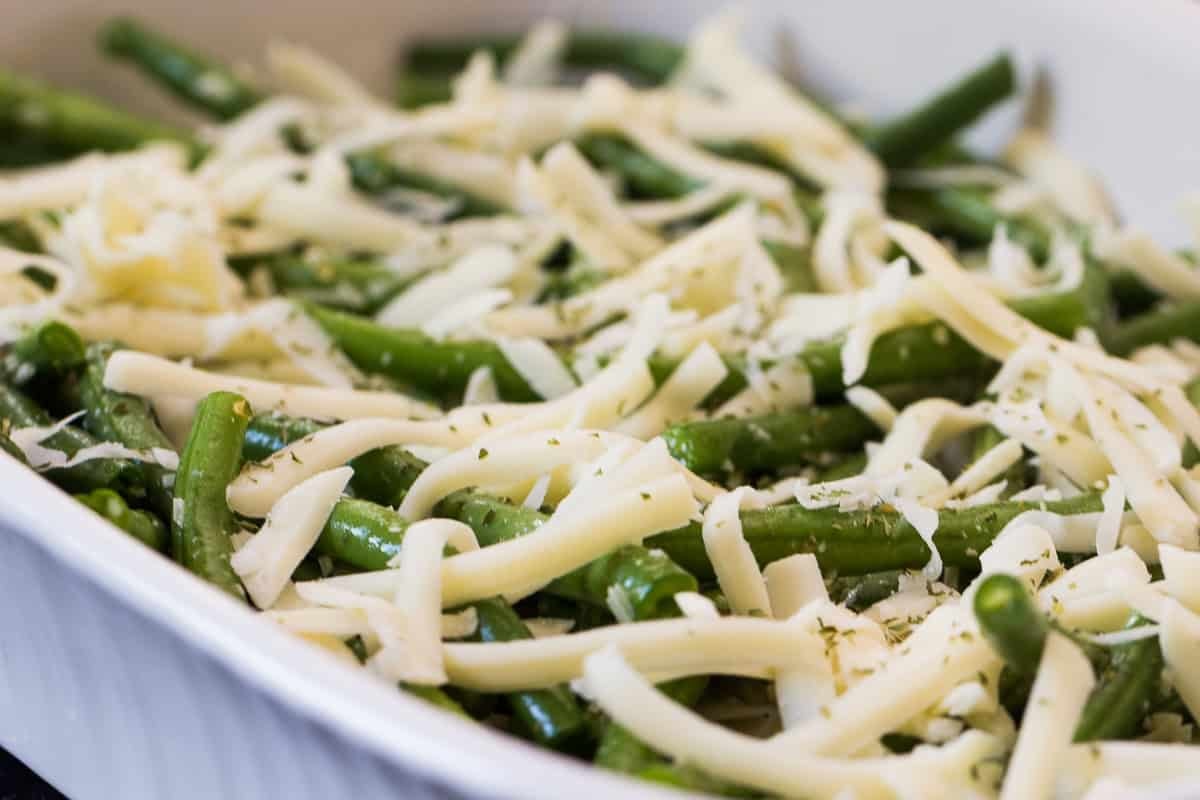 A close-up of green beans topped with shredded cheese and sprinkled with herbs, placed in a white baking dish.