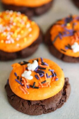 Close-up of chocolate cookies topped with orange frosting and colorful Halloween-themed sprinkles, featuring small ghost and bat shapes.