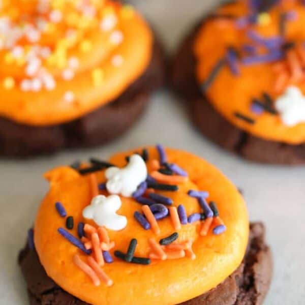 Close-up of chocolate cookies topped with orange frosting and colorful Halloween-themed sprinkles, featuring small ghost and bat shapes.