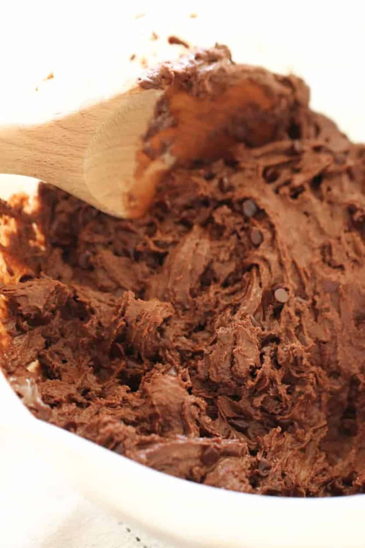 A wooden spoon stirs thick, chocolate cookie dough in a white mixing bowl.