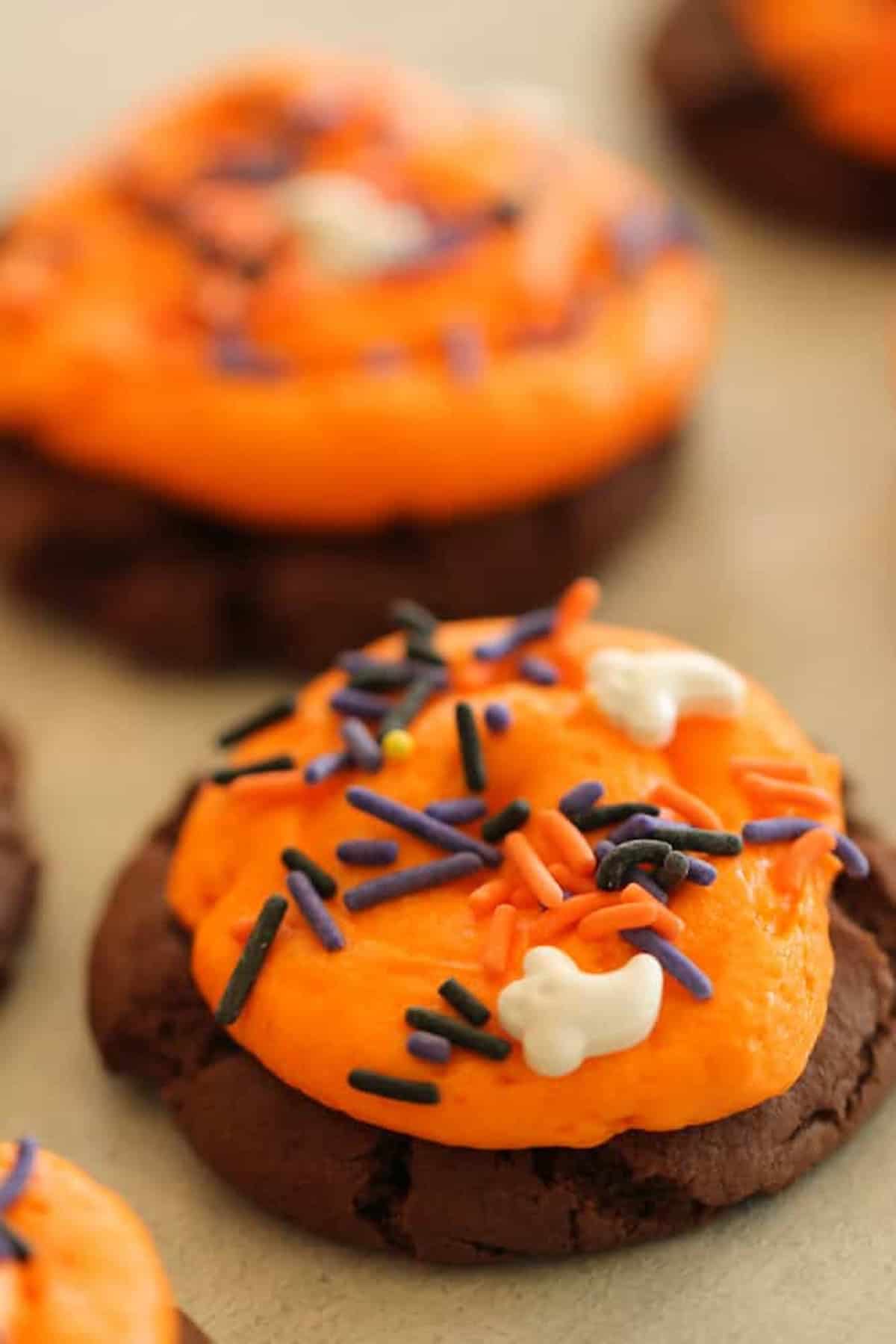 Close-up of chocolate cookies topped with orange icing and Halloween-themed sprinkles including bat and ghost shapes.