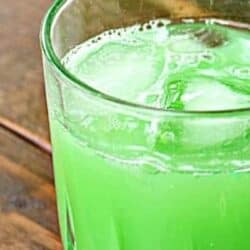 A close-up of a glass containing a green-colored beverage with ice cubes, placed on a wooden surface.