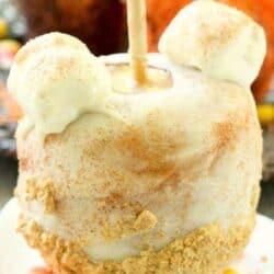 A caramel apple covered in white icing, dusted with cinnamon, topped with two marshmallows, and sprinkled with crushed cookies sits on a plate surrounded by candy corn.