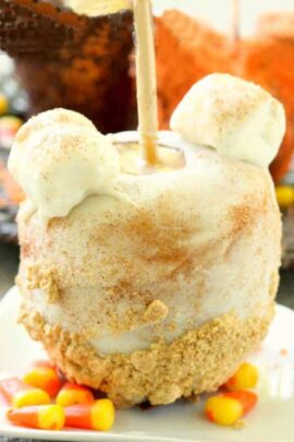 A caramel apple covered in white icing, dusted with cinnamon, topped with two marshmallows, and sprinkled with crushed cookies sits on a plate surrounded by candy corn.