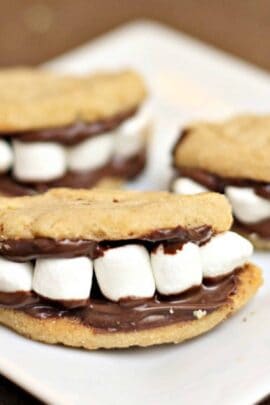 Three cookie sandwiches filled with marshmallows and chocolate, resembling smiling mouths with teeth, are placed on a white plate.