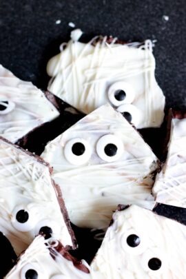 Pieces of chocolate bark coated in white icing and adorned with candy eyes, arranged on a black surface.