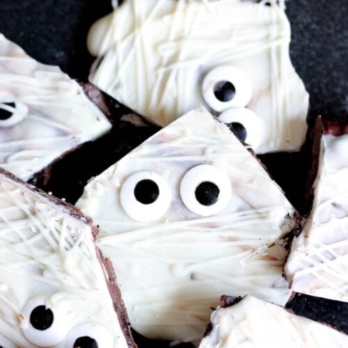 Pieces of chocolate bark coated in white icing and adorned with candy eyes, arranged on a black surface.