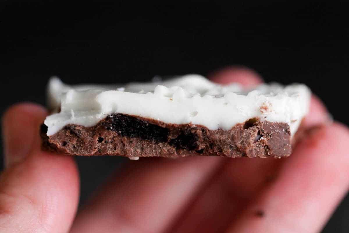 A close-up shot of a hand holding a small piece of chocolate with a white frosting layer on top.