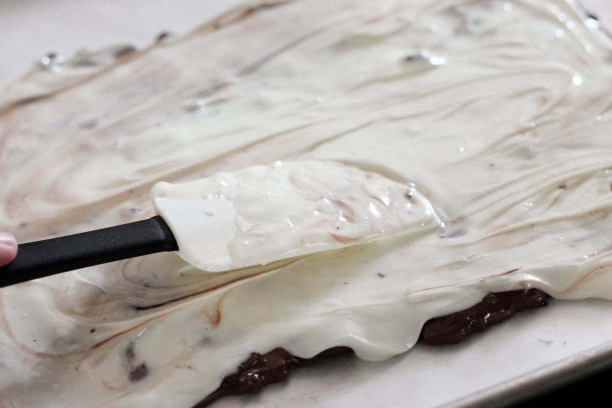 A spatula spreads melted white and dark chocolate on a flat surface, creating a swirl pattern on what appears to be a chocolate dessert base.