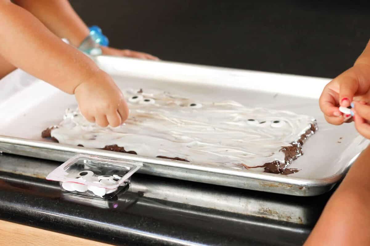 Two children are decorating a sheet of chocolate brownies with white frosting and adding candy eyes on top, using a baking sheet on a counter.