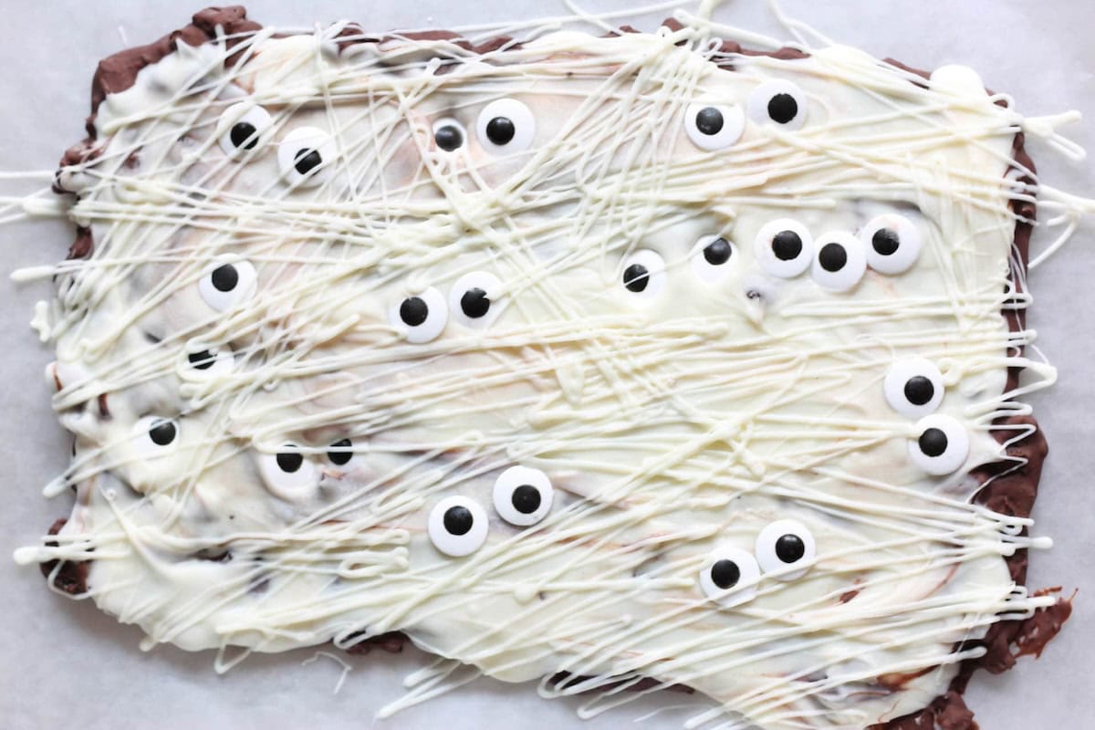 A rectangular chocolate bark decorated with white drizzles and small candy eyes, resembling a mummy.