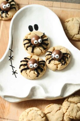 Close-up of three spider-themed cookies on a ghost-shaped plate. The cookies are decorated with candy eyes and chocolate bodies on a wooden surface, with additional cookies scattered around.