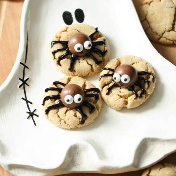 Close-up of three spider-themed cookies on a ghost-shaped plate. The cookies are decorated with candy eyes and chocolate bodies on a wooden surface, with additional cookies scattered around.