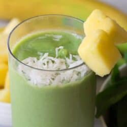 A glass of green smoothie topped with shredded coconut and garnished with two chunks of pineapple on the rim, with a partial view of a banana and a bowl of pineapple chunks in the background.