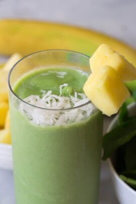 A glass of green smoothie topped with shredded coconut and garnished with two chunks of pineapple on the rim, with a partial view of a banana and a bowl of pineapple chunks in the background.