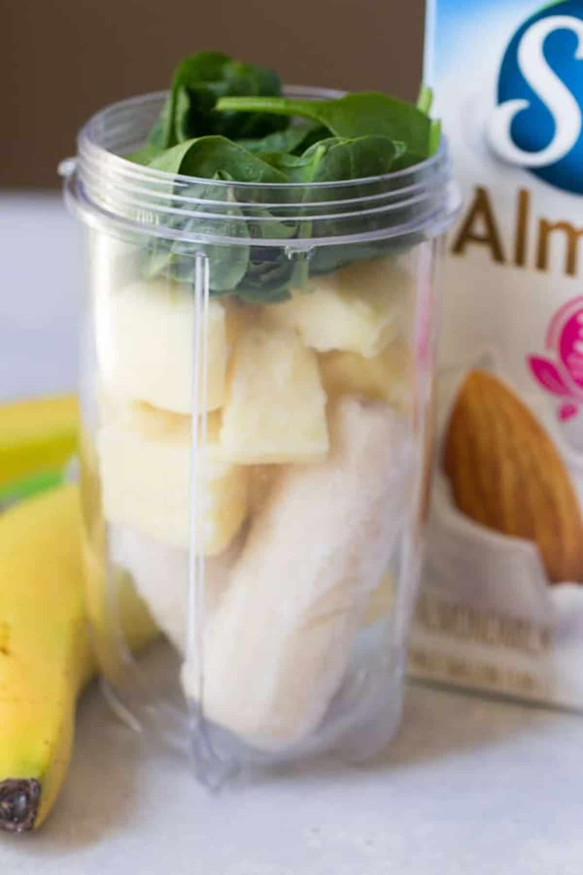 A blender cup filled with spinach, pineapple, and banana is placed next to a banana and a carton of almond milk.
