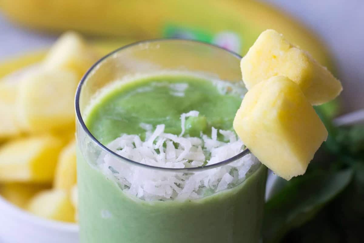 A close-up of a green smoothie topped with shredded coconut and two pineapple chunks on the glass rim. A blurred background shows more pineapple pieces and a banana.