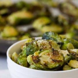 A white bowl filled with roasted Brussels sprouts, sprinkled with grated cheese, and a blurred background with more Brussels sprouts.