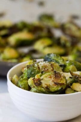 A white bowl filled with roasted Brussels sprouts, sprinkled with grated cheese, and a blurred background with more Brussels sprouts.